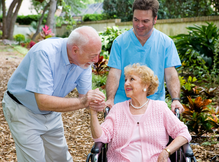 Senior husband visiting his disabled wife on the grounds of the nursing home while a nurse pushes her wheelchair.